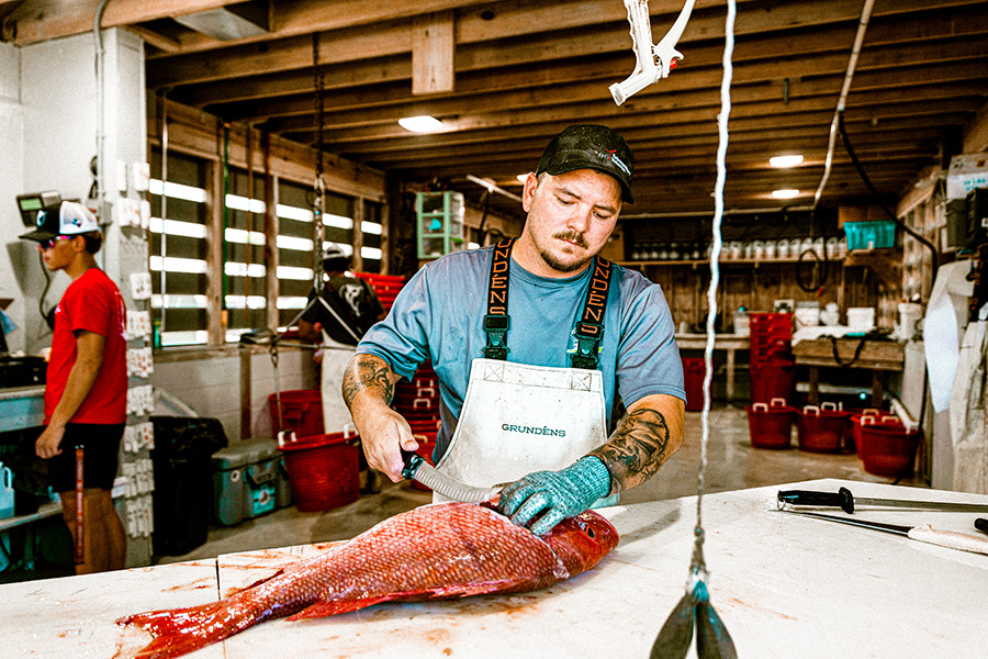 A fisherman in overalls and gloves cleaning a large red fish on a white table at a busy fish cleaning station, with another person working in the background.