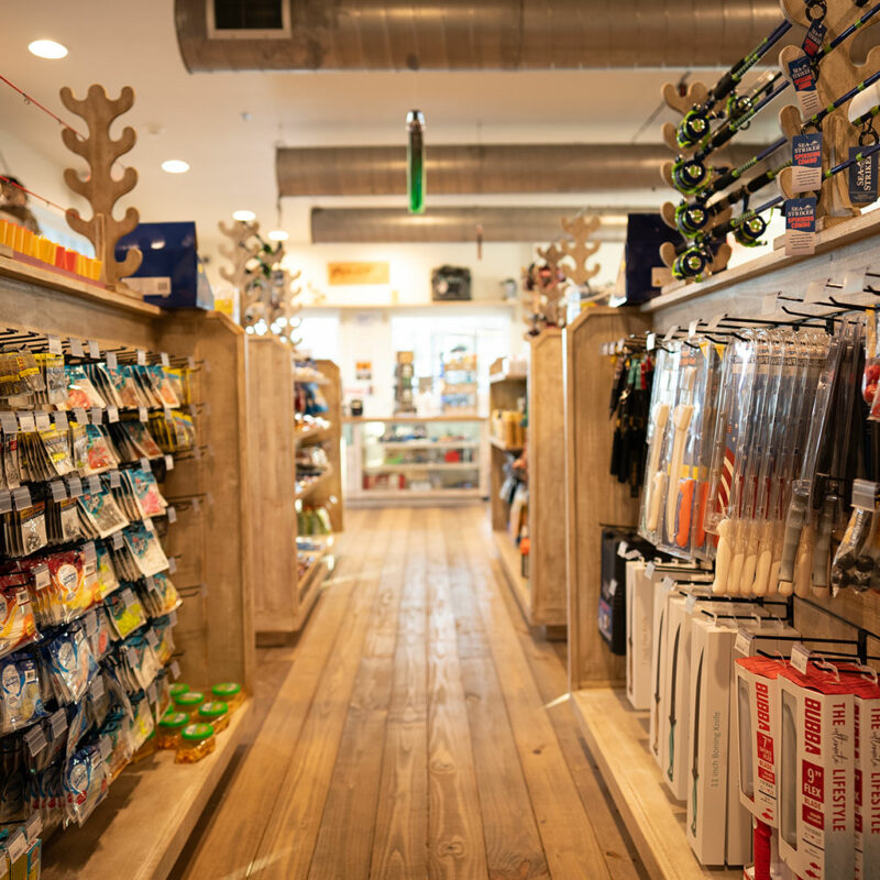 Aisle view inside a fishing supply store with wooden shelves stocked with various fishing lures, tackle, and accessories, featuring wooden floor and maritime-themed decorations.