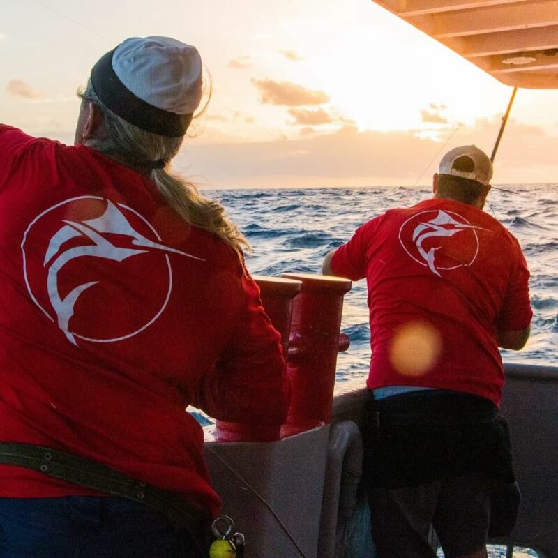 Two fishermen in red shirts with a marlin logo on the back, fishing on a boat at sunrise on the open ocean.