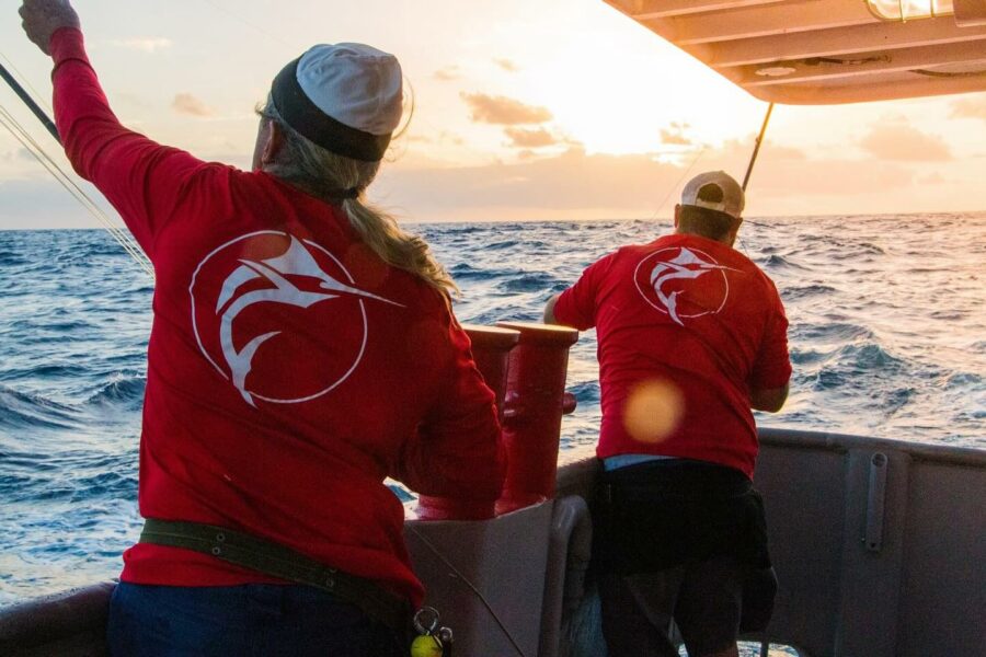 Two fishermen in red shirts with a marlin logo on the back, fishing on a boat at sunrise on the open ocean.